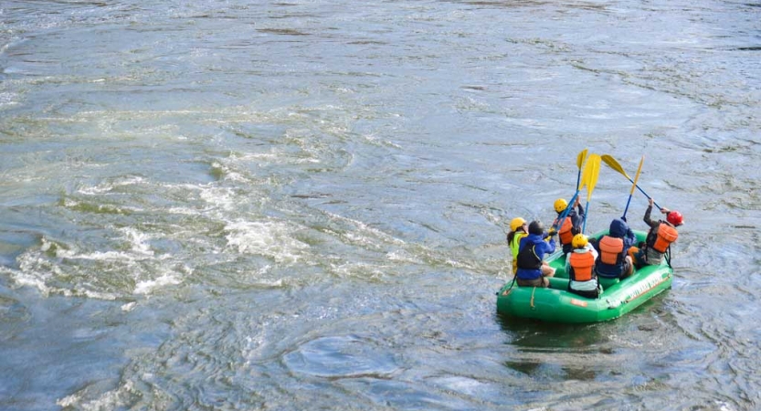 A group of students wearing safety gear sit in a raft and raise their paddles into the air.